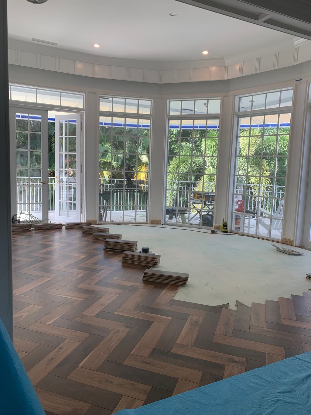 Herringbone wood flooring being laid across a curved bay, with stacks of planks waiting by the French doors.