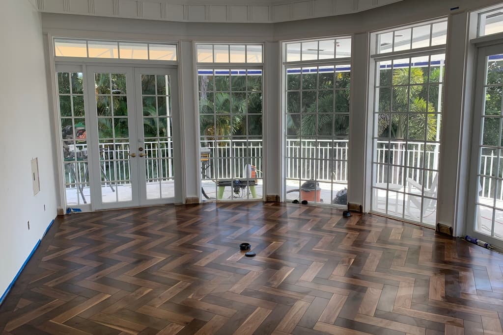 A curved bay of French doors and windows opens to a terrace and palms, above a newly finished herringbone wood floor.