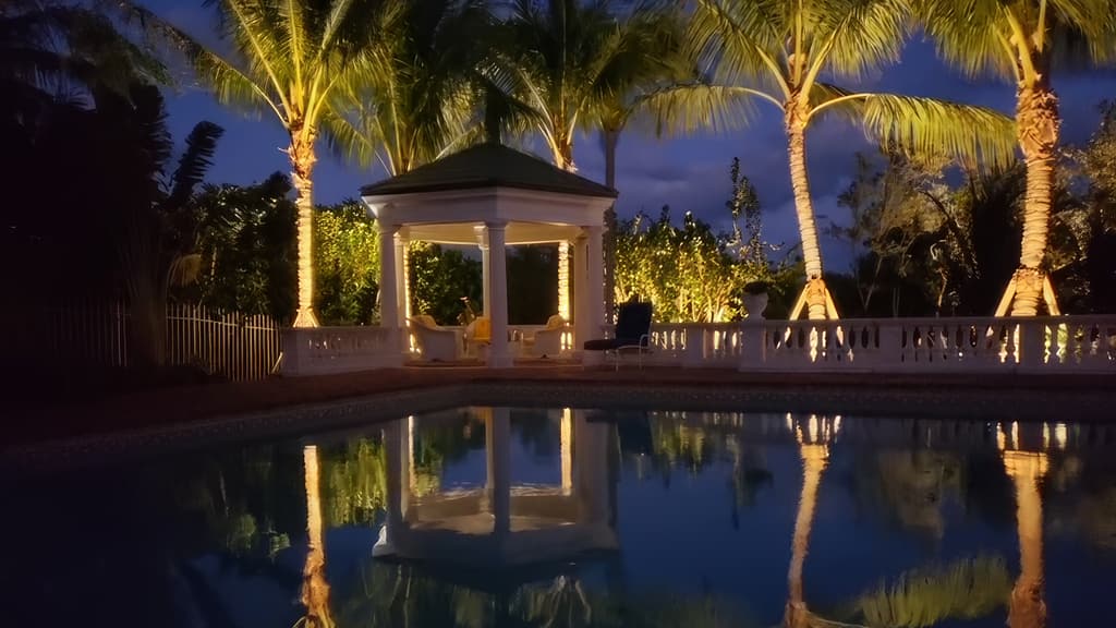 A pool at night mirroring a lit gazebo and a row of uplit palm trees.